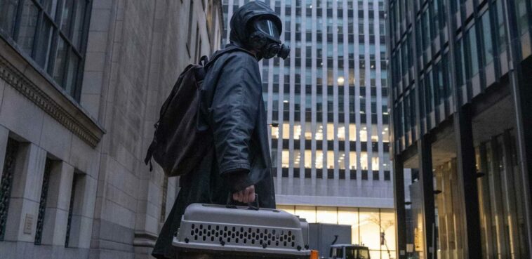 Yorick, in full black hazmat suit and gas mask, carries a pet carrier through a city street and looks up at the sky