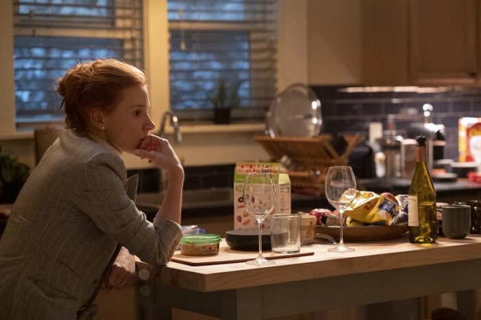 In this image from Scenes from a Marriage, Mira (Jessica Chastain) is depicted seated, wearing a business suit, in her and Jonathan's dimly lit kitchen, with her face resting on her hand, looking pensively to the right.