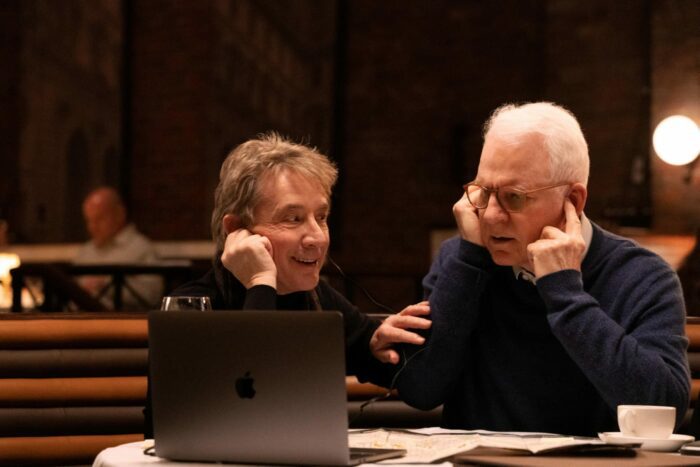 Oliver (Martin Short) and Charles (Steve Martin) sit at a table in a dark restaurant with their hands to their ears listening to a podcast that is playing on Oliver's computer which is in the foreground on the table