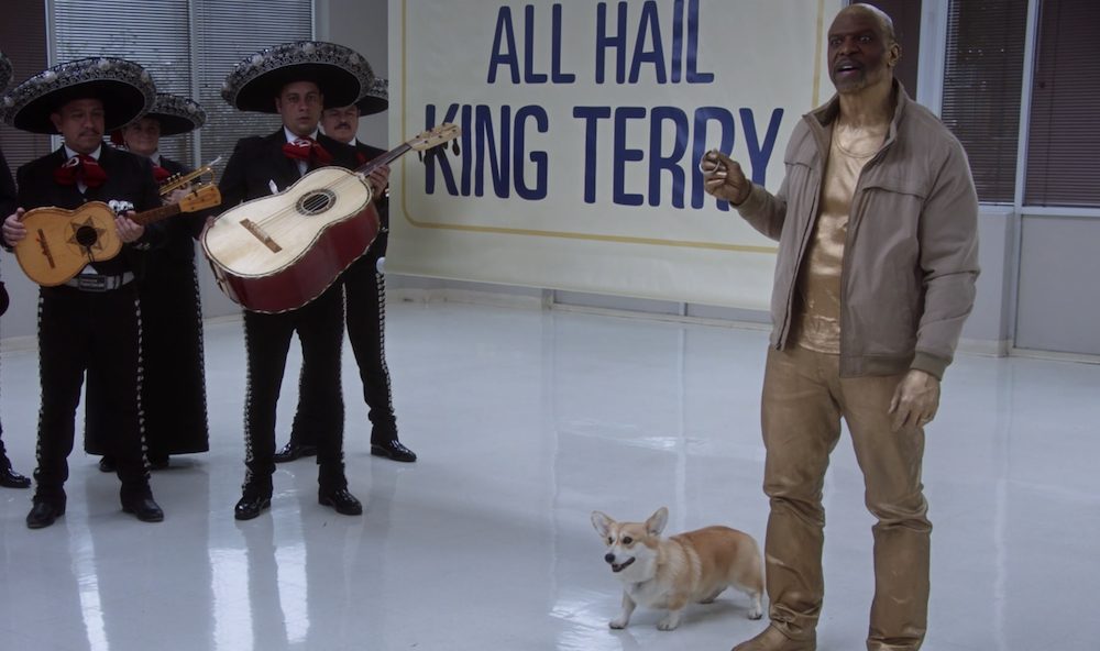 Terry dressed in gold with a banner that reads 'All Hail King Terry' behind him, Cheddar the dog by his side and a mariachi band in the background