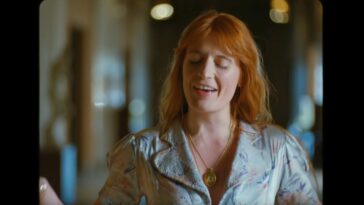 Florence Welch closes her eyes as she stands in a hallway and sings in the music video for "Hunger" by Florence + The Machine