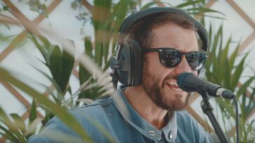 Bearded singer in dark sunglasses (Grant Nicholas of Welsh rock band Feeder) singing to the mic on a leafy background