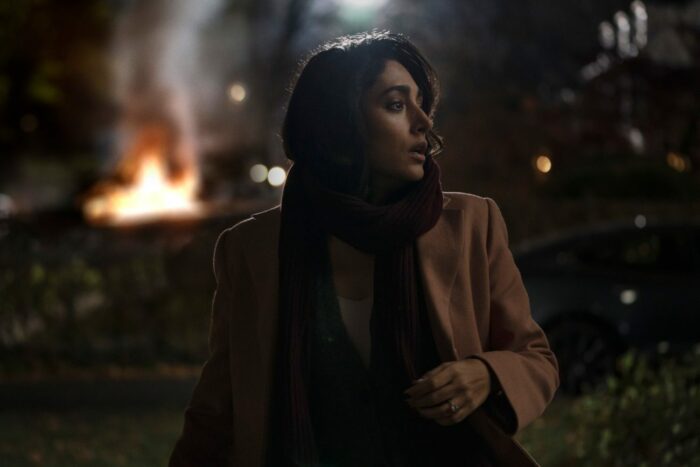 Aneesha looks back over her shoulder, as cars burn in the background