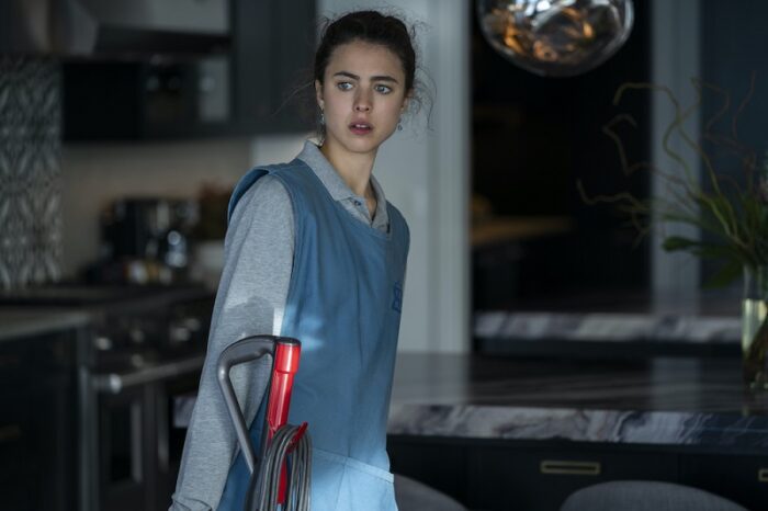 Margaret Qualley as Alex is depcted in a maid's apron and carrying a vacuum cleaner in the kitchen of a well-appointed home in this image from Netxlix's limited series Maid.