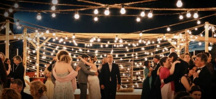 Kevin stands alone, with bright lights strung all around him, while others dance at a wedding