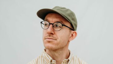A man with circular glasses, a light brown collared shirt, and a faded green cap stares away from the camera, his head turned to his right (the image is of the singer-songwriter Cuchulain)