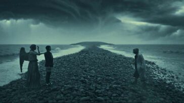 The Rogue Angel stands to the side with Claire (Anabel Scholey) looking accross a dark inlet by the ocean to the Doctor (Jodie Whittaker) the entire image is grey and far away