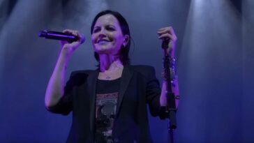 A woman wearing a black blazer, black t-shirt, and silver bangles smiles and holds a microphone on stage (Dolores O'Riordan of The Cranberries)