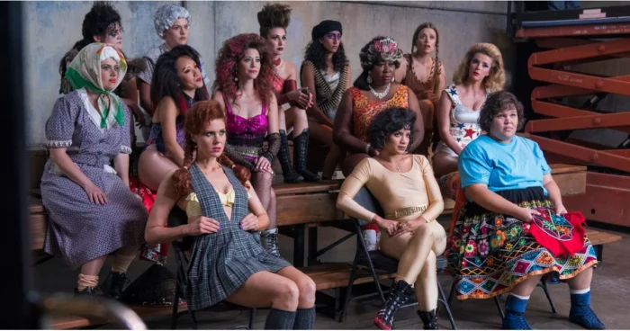The cast of GLOW sitting on gym benches. 