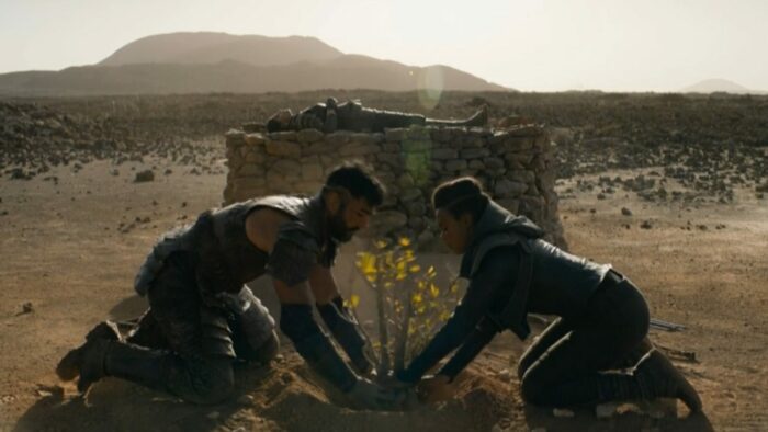 Rowan and Salvor lower a tree sappling into a hole, Phara's body lies behind them on a stone dias