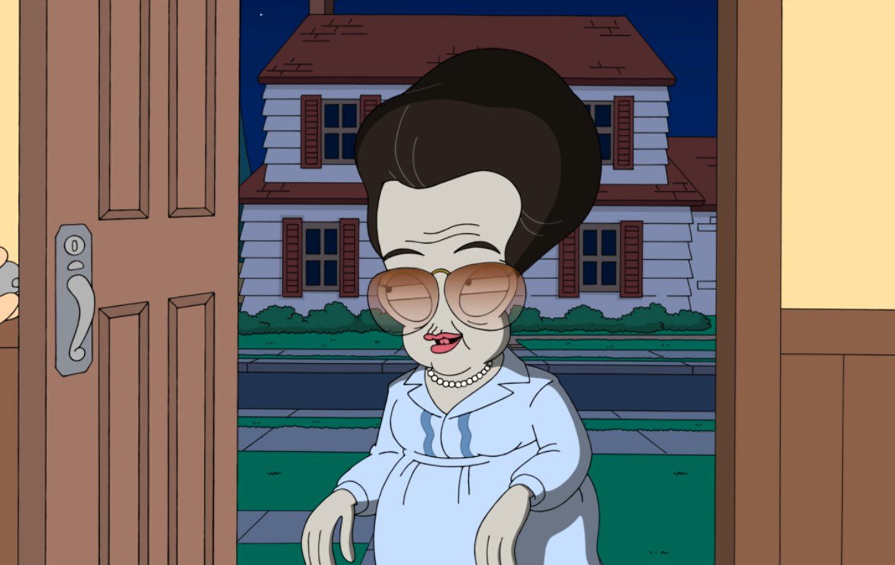 A bulbous-headed grey alien dressed as a woman in a blue dress wears tinted aviator glasses, a pearl necklace, and a poofy grey wig; she stands in the doorway of a suburban house (Roger as Ruby Zeldastein in American Dad!)