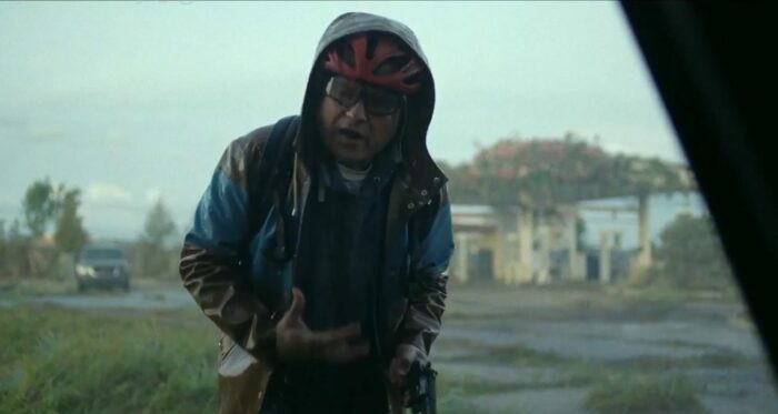 Brian (Enrico Colantoni) standing in the rain wearing a bicycle helmet holding a gun