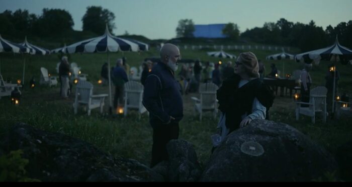 Gil (David Cross) and The Conductor (Lori Petty) talk in the shadows in front of umbrellas and people set up to watch the show in Station Eleven Episode 4