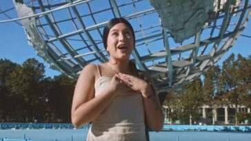 A young woman with black hair tied back in a ponytail wears a beige tanktop and stands in front of a metallic globe sculpture on a sunny day (KAYA in the Harana x Half Free music video)