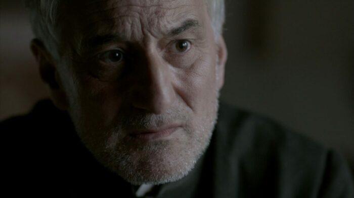 A man with a thin grey beard, brown eyes, and tan skin in a priest's frock wears a concerned expression.