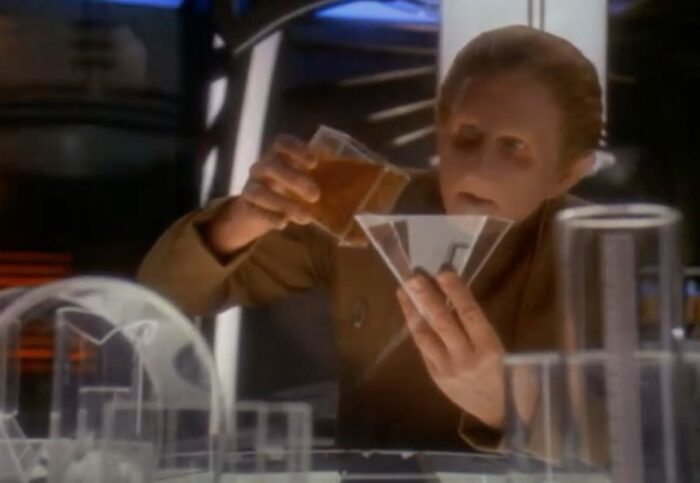 Odo pours some amber liquid from a square glass beaker into a triangular one, looking very focused on his experiment.