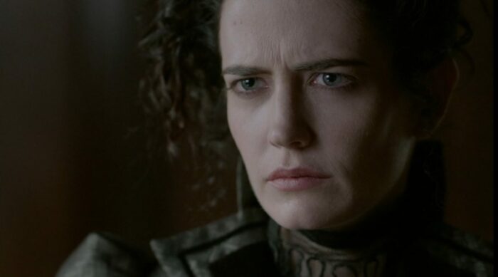 A woman with pale white skin, blue eyes, and dark brown hair wears a serious expression with a furrowed brow.