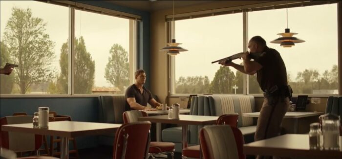 Jack Reacher in a t-shirt sits at a diner table with a coffee cup in front of him. A police offers stands in front of him with a shotgun pointed at him. 