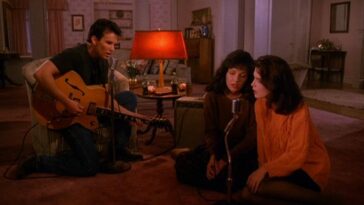 James kneels with a guitar in a living room as Maddy and Donna sit on the floor