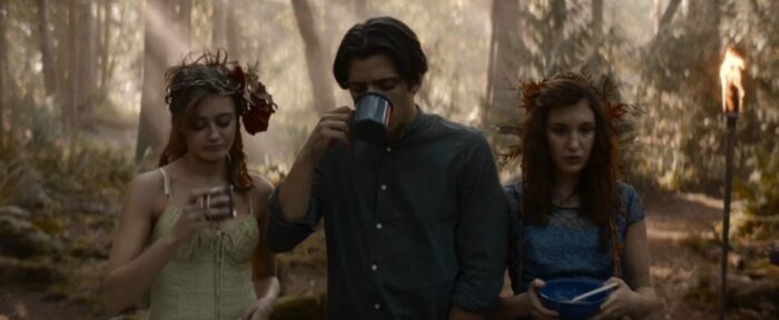 Travis takes a drink from a metal cup as Jackie and Shauna stand to each side of him in the woods in 1996 in Yellowjackets "Doomcoming"