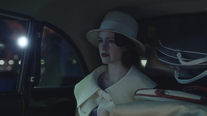 Midge rides home from the airport in the backseat of a cab, in her fancy white outfit and hat