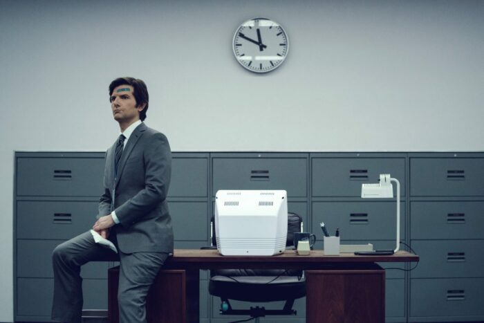 Mark sits against a desktop, with a clock on the wall behind him
