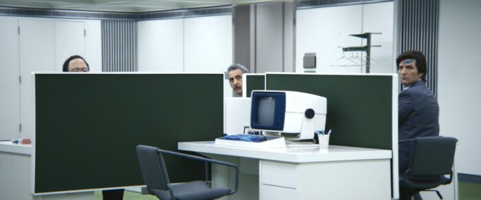 Irv peeks out from behind a desk partition, with Mark to the right side of the frame, and a computer in view to the front