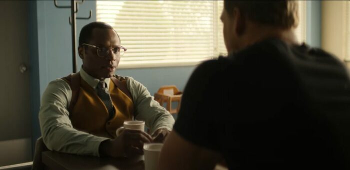 Finley sits in a tie and vest in a diner while talking with Jack Reacher over a cup of coffee