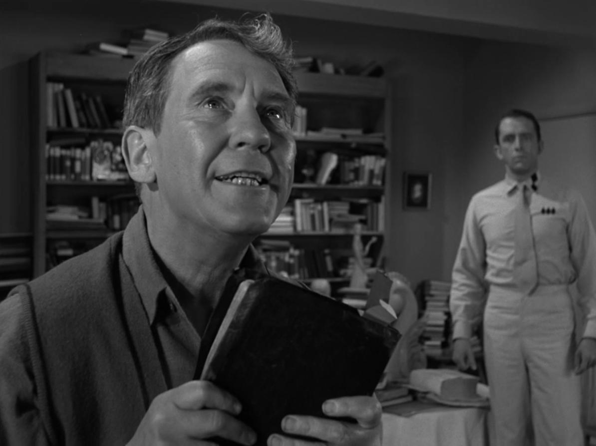 Burgess Meredith as Romney Wordsworth from "The Obsolete Man", front, holding a book and looking upwards. Fritz Weaving as The Chancellor standing in the background, looking at him. Behind them both is a wall lined with books