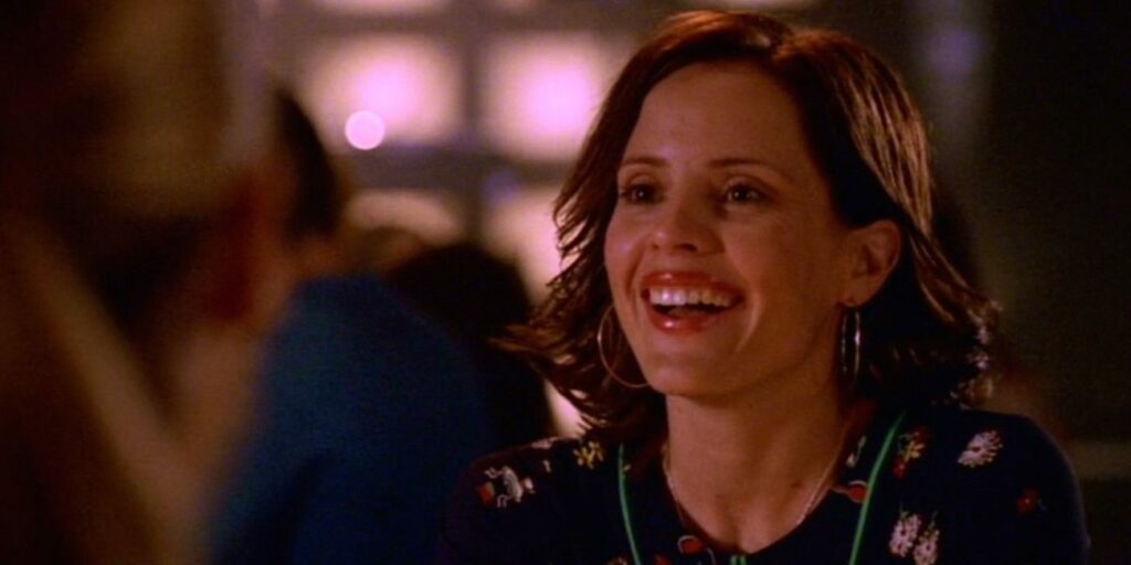 Anya Jenkins (Emma Caulfield), wearing a black, floral-print shirt and large hoop earrings, looks ahead smiling..