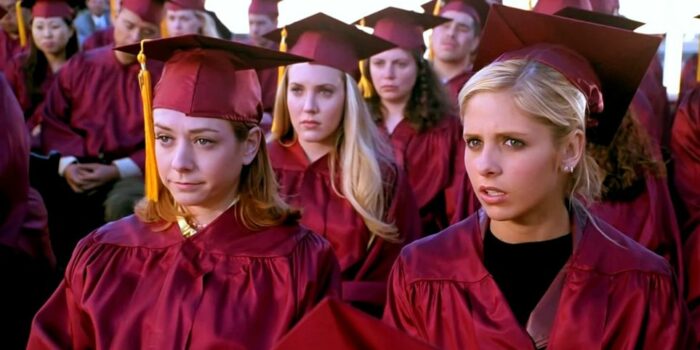 Buffy and Willow look on from the stands in graduation robes, surrounded by other students