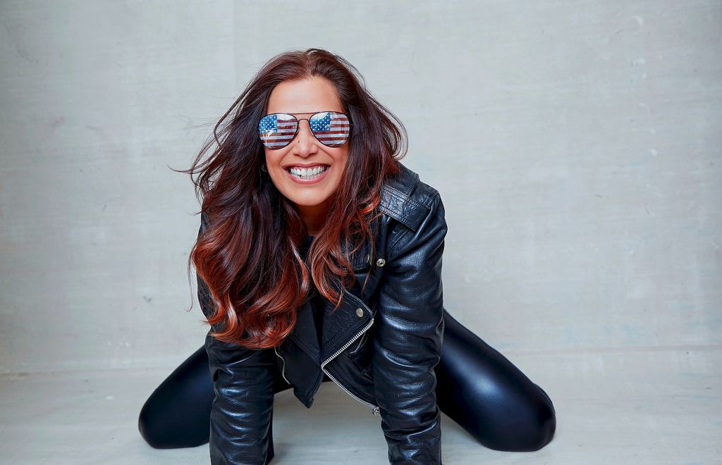 Actress Kamala Lopez wearing American flag sunglasses smiles while kneeling on the ground