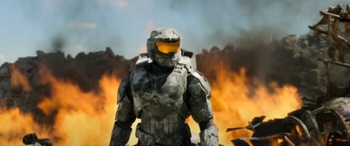 Pablo Schreiber as The Master Chief in green armor standing with his back to a raging fire