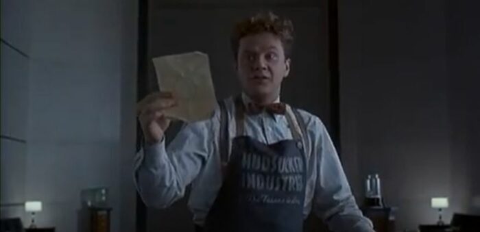 Tim Robbins wearing an apron that reads "Hudsucker Industries" holds up a piece of paper with a circle on it