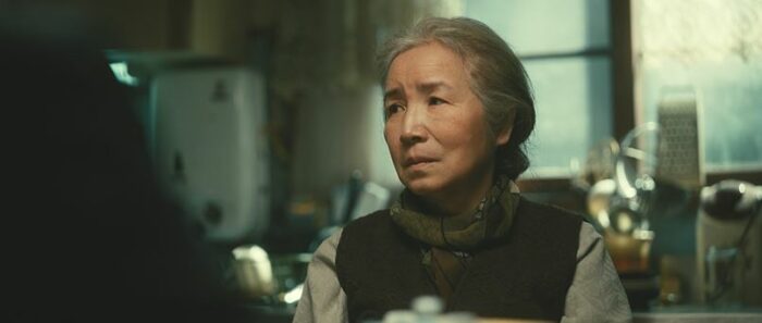 An older Korean woman looks on, sitting in a kitchen