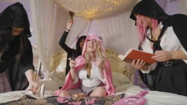 Scene Queen sits on a bed surrounded by others holding books and other objects, in the video for Pink Rover