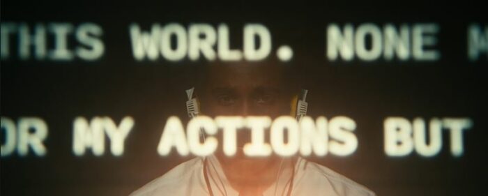 Milchick sits wearing headphones, words on a screen in front of his face read, in fragments, "this world. none...my actions but"