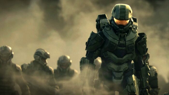 Master Chief walks in front of a group of marines on a dusty road.