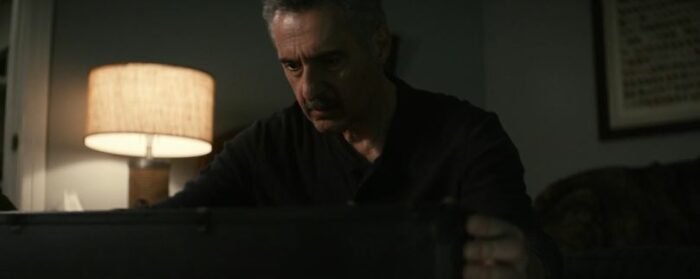 Irv, in a black shirt, leans over a chest, with a lamp in the background