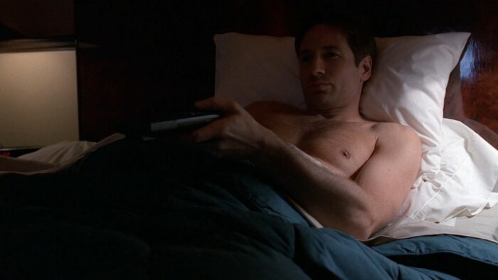 Agent Mulder lies naked in bed holding a TV remote
