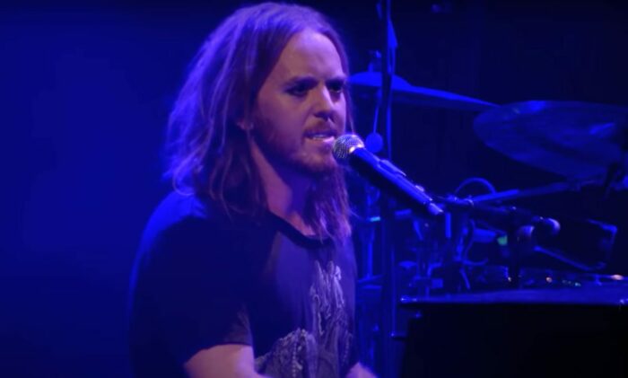 Tim Minchin, bathed in blue light, wears a T-shirt as he sings into a microphone