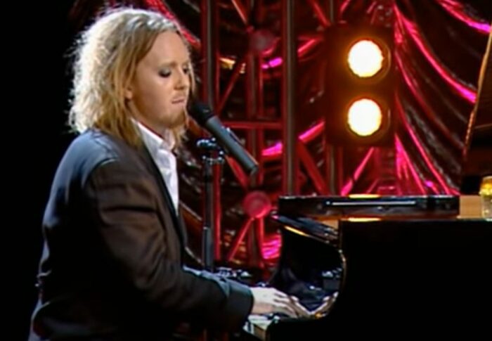 Tim Minchin, with eyes downturned, sits in front of a piano