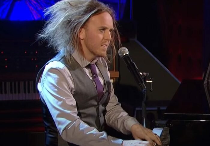 Tim Minchin, in a vest and tie, sits in front of a piano
