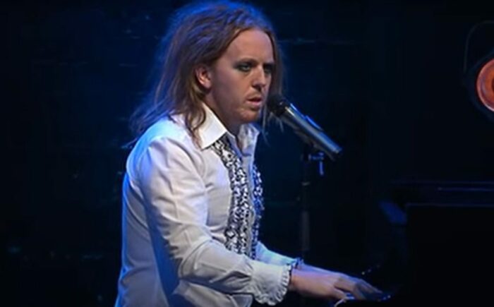 Tim Minchin in a white frilly shirt, in front of a piano and a microphone