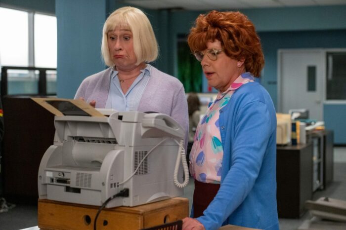 Kathy and Cathy stand looking at a fax machine, bemused