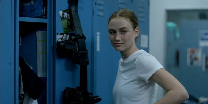 Maddie Bosch looks to the left as she stands in a white tshirt in front of blue lockers, where her gun belt hangs. 