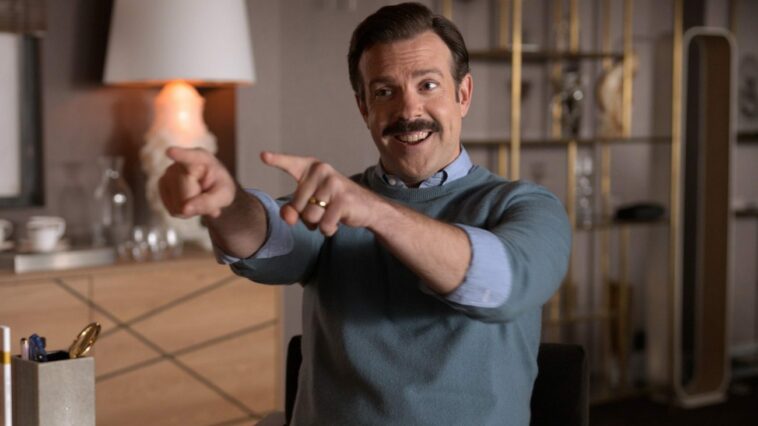 Ted Lasso is playfully pointing both of his index fingers over a desk