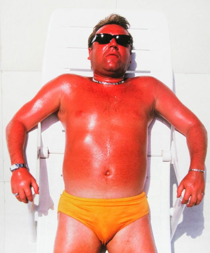 A tan man in a speedo and sunglasses lies on a raft in a pool
