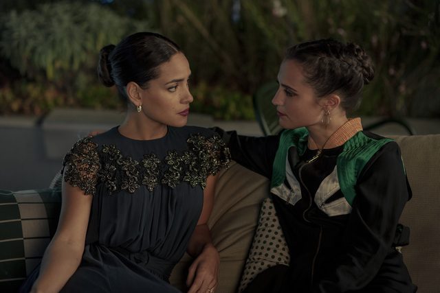 Laurie (Adria Arjona, left) and Mira (Alicia Vikander) share an embrace on an outdoor patio at night.