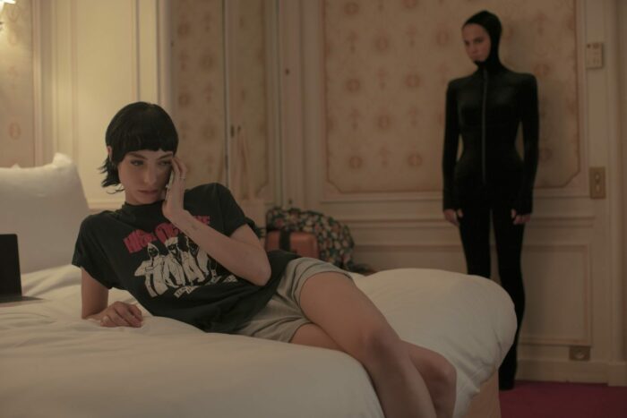 Image from Irma Vep: Regina (Devon Ross) sits on a bed as Mira (Alicia Vikander) watches in her catusuit.